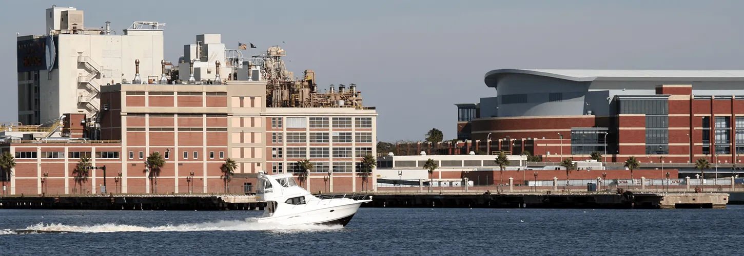 White motorboat speeding along a harbor with industrial buildings and a modern stadium in the background.